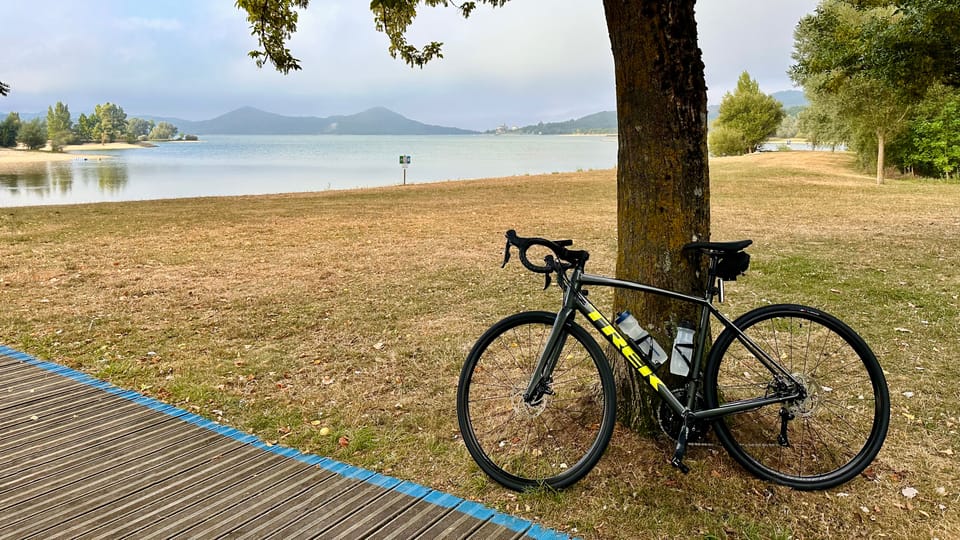 A road bike leaning against a tree on a grassy shore, with a calm reservoir and distant hills under a cloudy sky in Vitoria.
